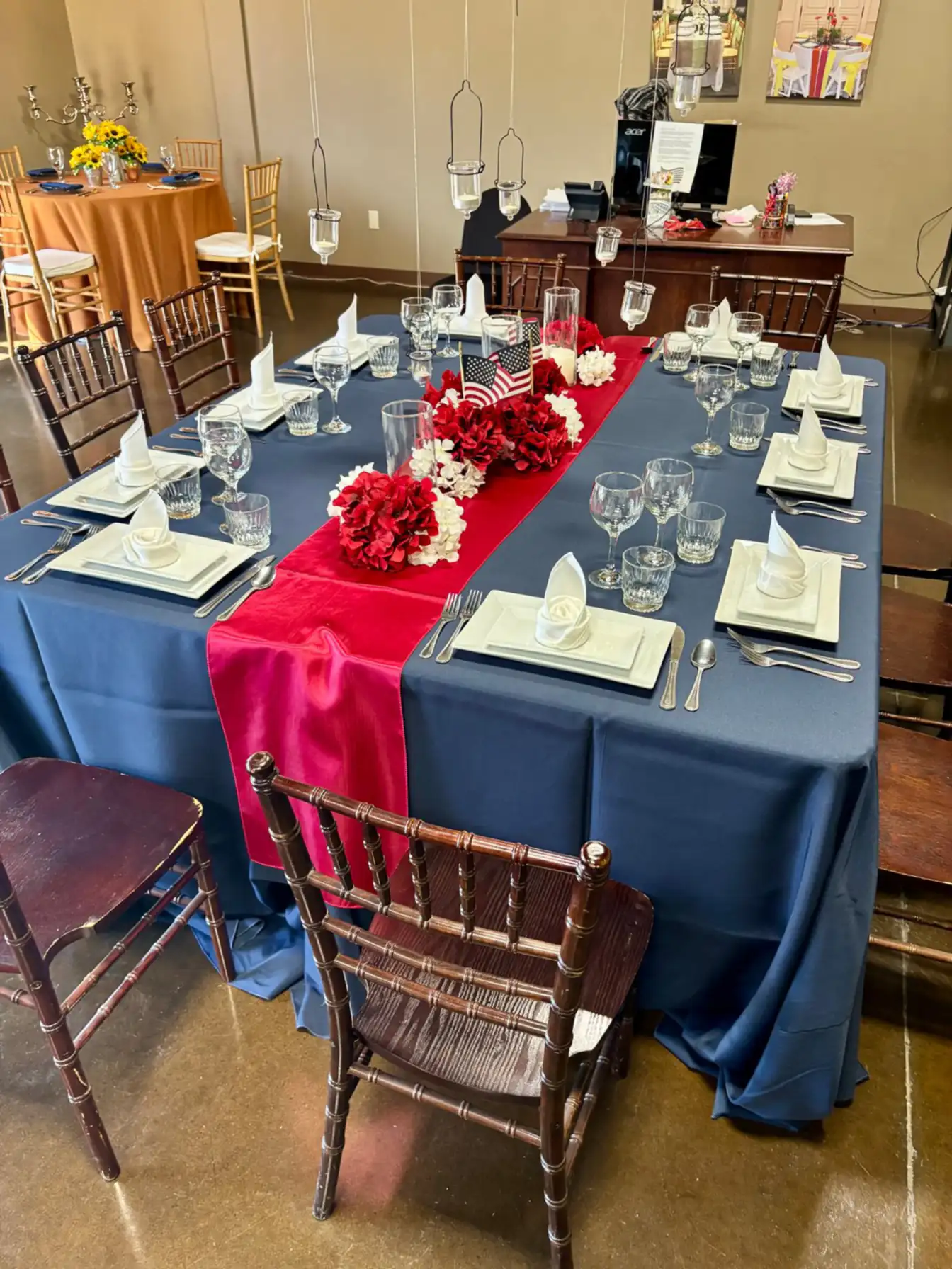 Square dinner table in navy linen with a red satin runner, rose-folded white napkins, small American flags, and red and white hydrangea florals